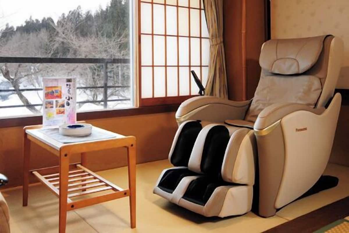 Una habitación con sillón de masajes, una mesita con una revista y una ventana con vistas a árboles.