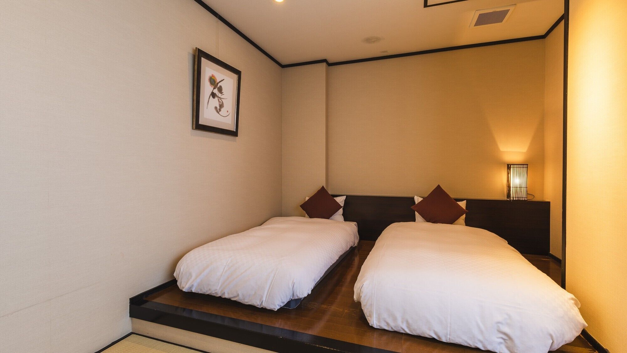 A hotel room with two single beds, a framed artwork on the wall, and a bedside lamp.