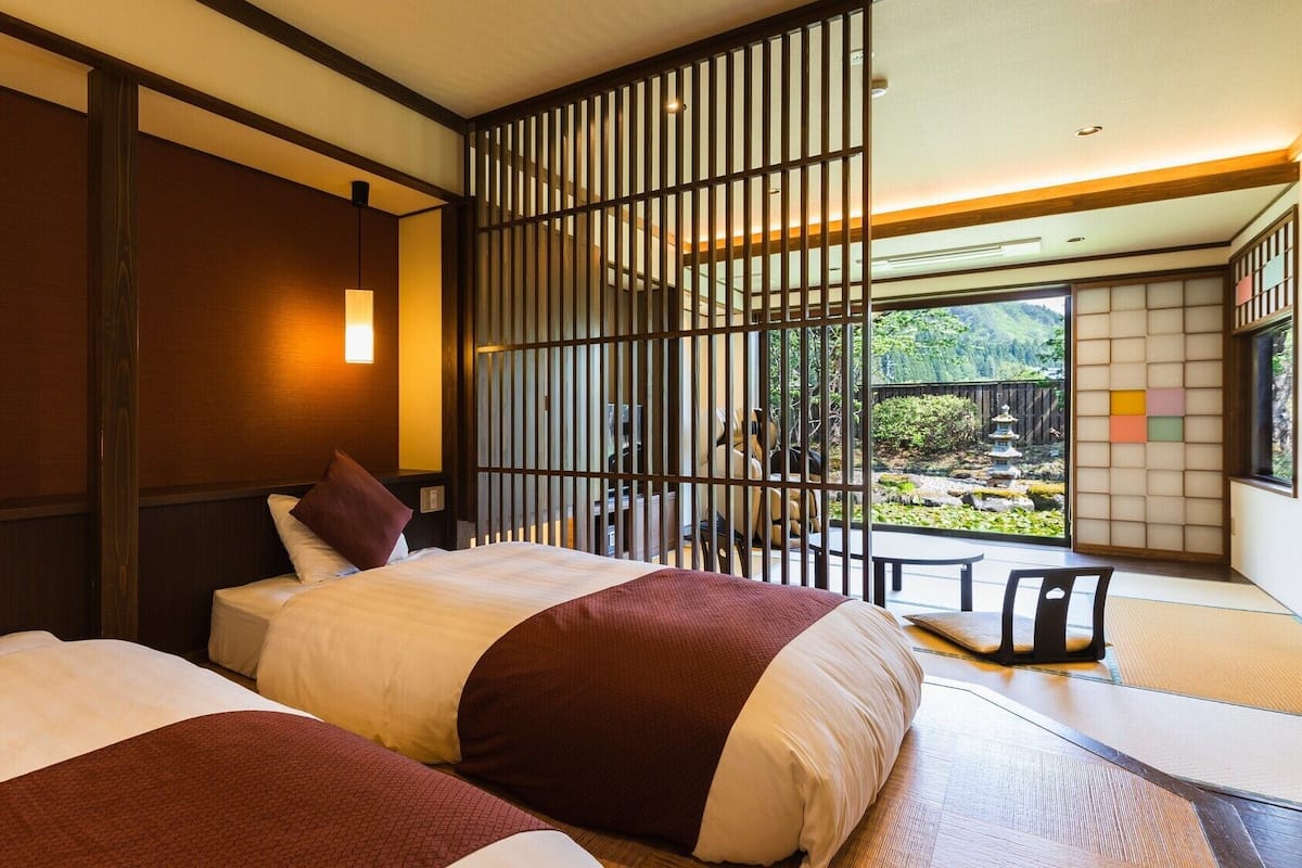 Una habitación tradicional japonesa con suelo de tatami, dos camas con ropa de cama roja y blanca, una puerta corredera y vistas a un jardín con una pagoda.