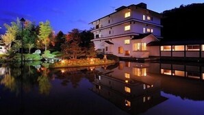 A multi-story building with a reflective water body in front, surrounded by trees and a clear sky. - Kajikaso (Yonezawa)