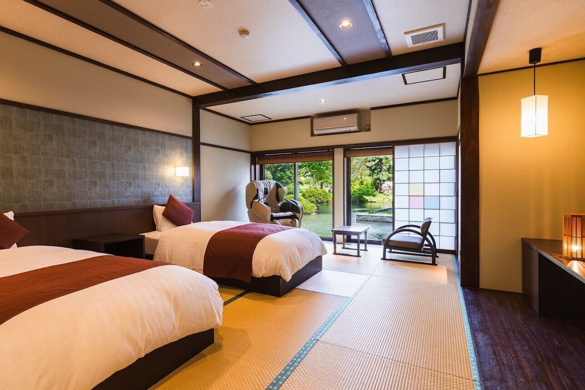 Habitación de hotel con dos camas, una mesita, una silla y vistas al exterior.