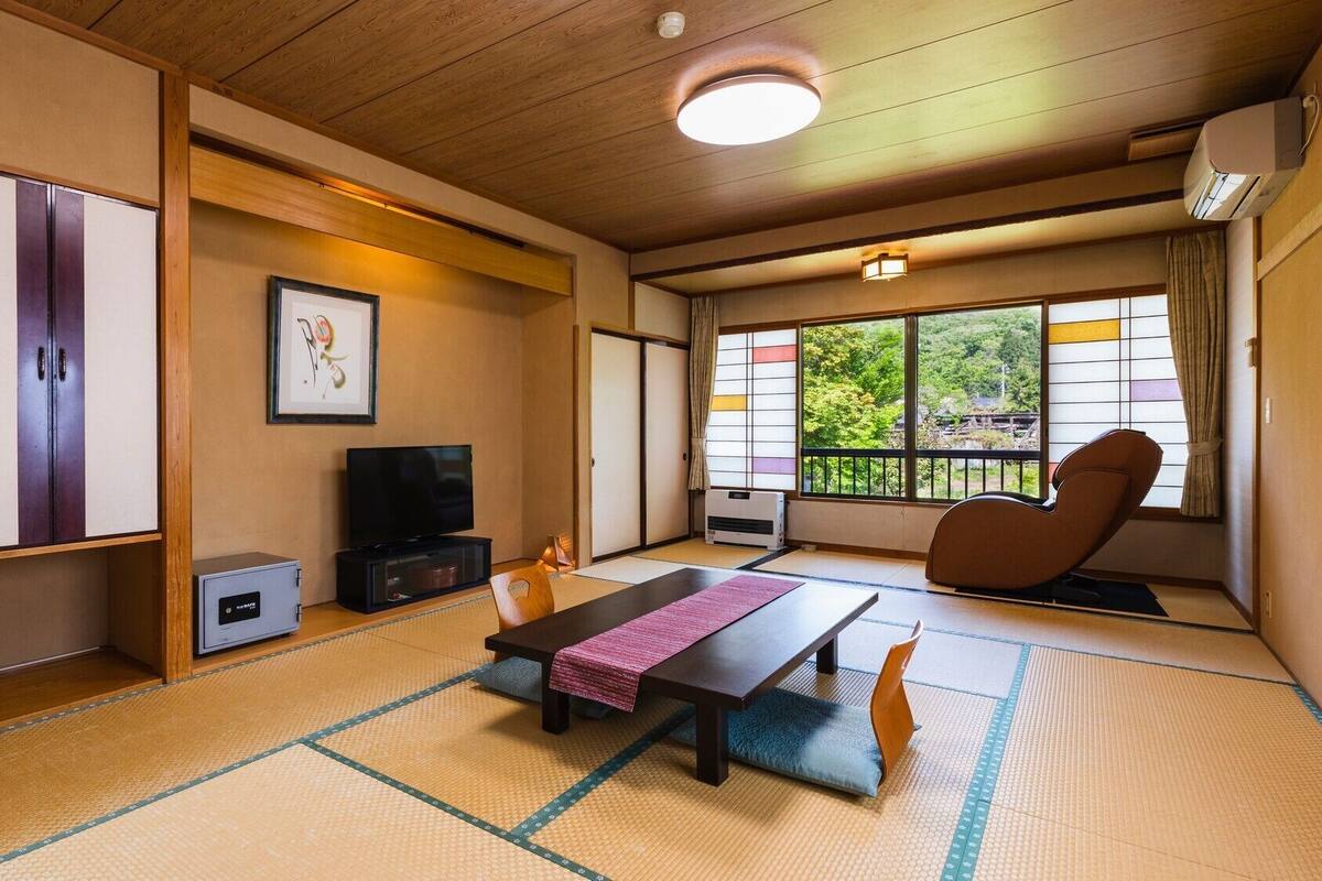 Una habitación japonesa tradicional con suelo de tatami, televisor, futón y una puerta corredera que da a un balcón con vistas.