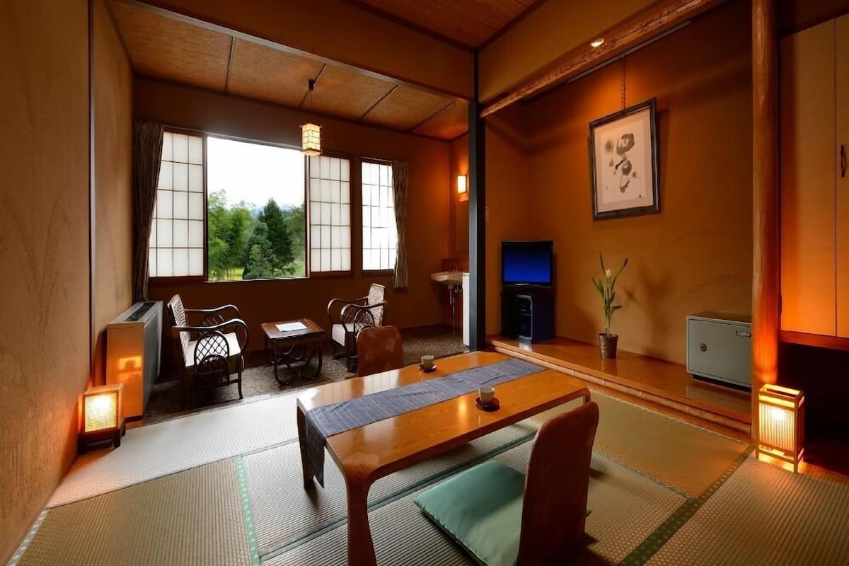 Una habitación japonesa tradicional con suelo de tatami, una mesita baja, sillas, un televisor y vistas a zonas verdes.