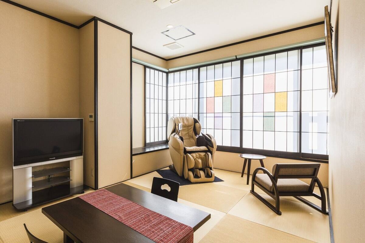 Una habitación con suelo de tatami, televisión, sillón de masaje, una mesita y una ventana con vistas.