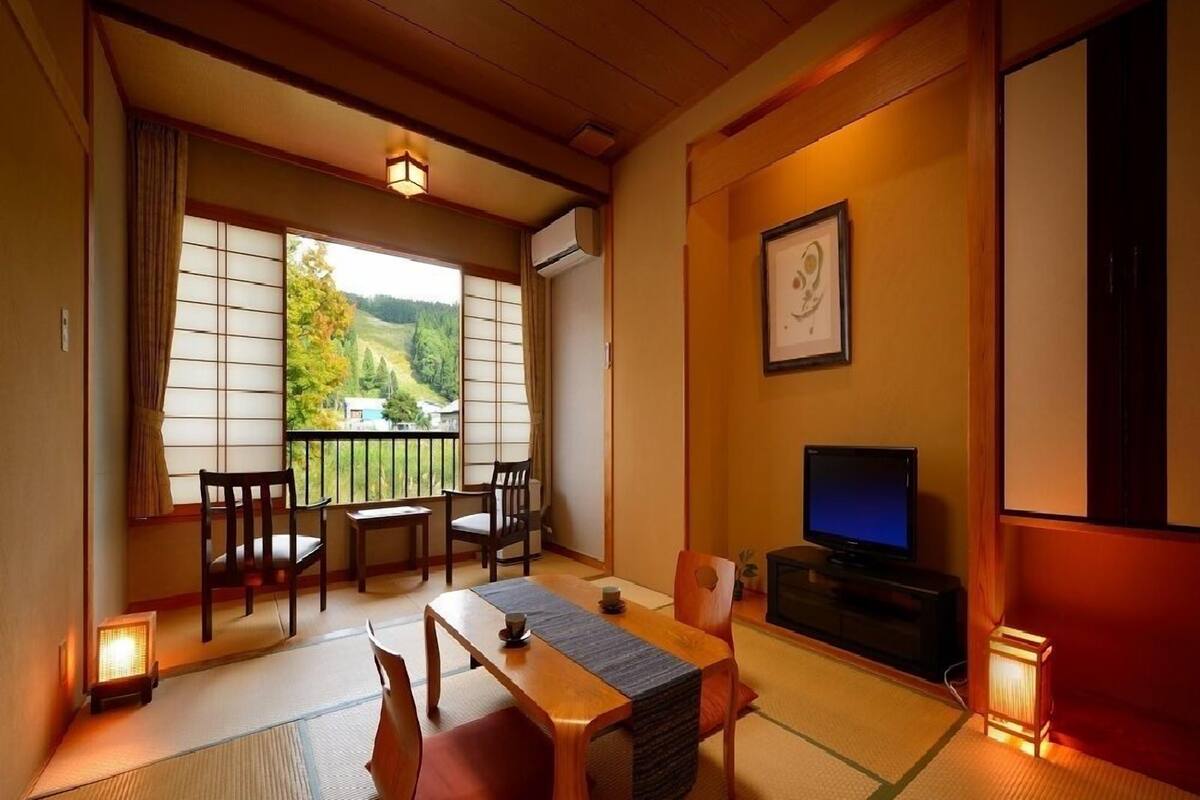 Una habitación japonesa tradicional con suelo de tatami, una mesa baja, sillas, un televisor y vistas a un paisaje verde.