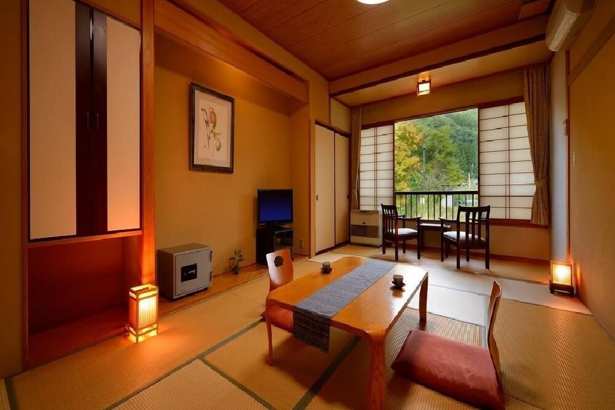 Una habitación japonesa tradicional con suelo de tatami, una mesa baja, sillas, un televisor y una ventana con vistas a árboles.