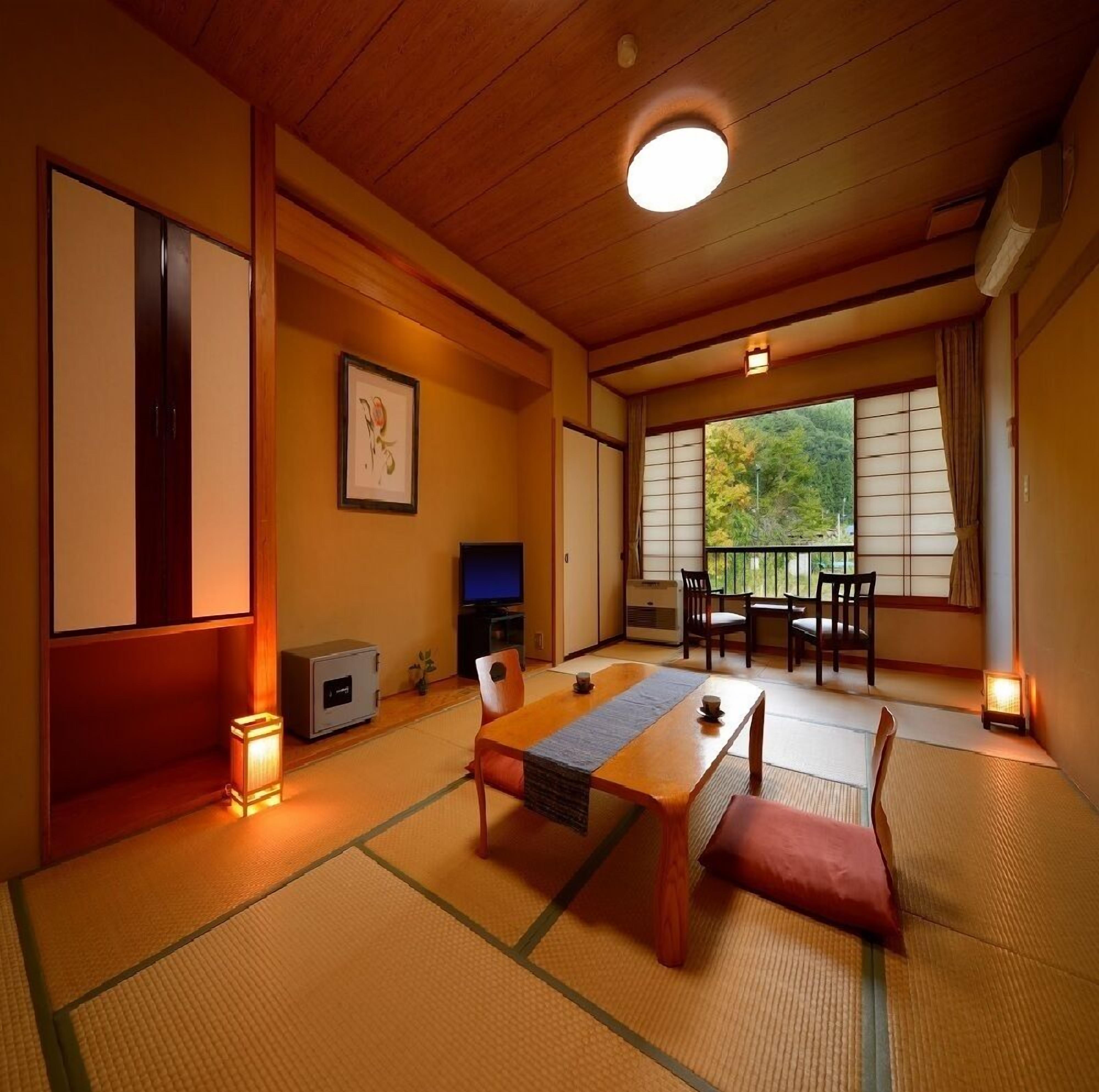 A traditional Japanese room with tatami flooring, a low table, chairs, a television, and a sliding door leading to a balcony.