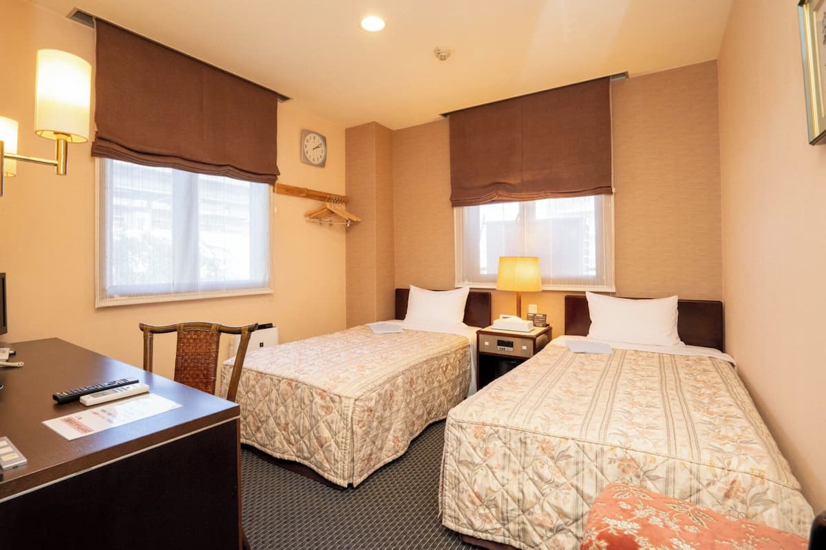 A hotel room with two beds, a desk, a chair, a clock, and a window with curtains.