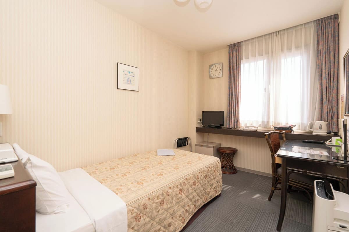 A hotel room with a bed, a desk with a computer, a chair, a clock, and a window with curtains.