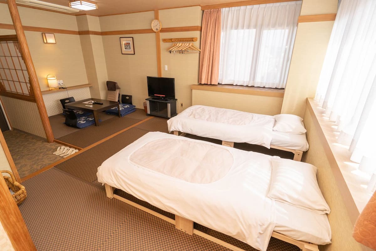 A hotel room with two beds, a television, a desk with a chair, a clock, and a window with curtains.
