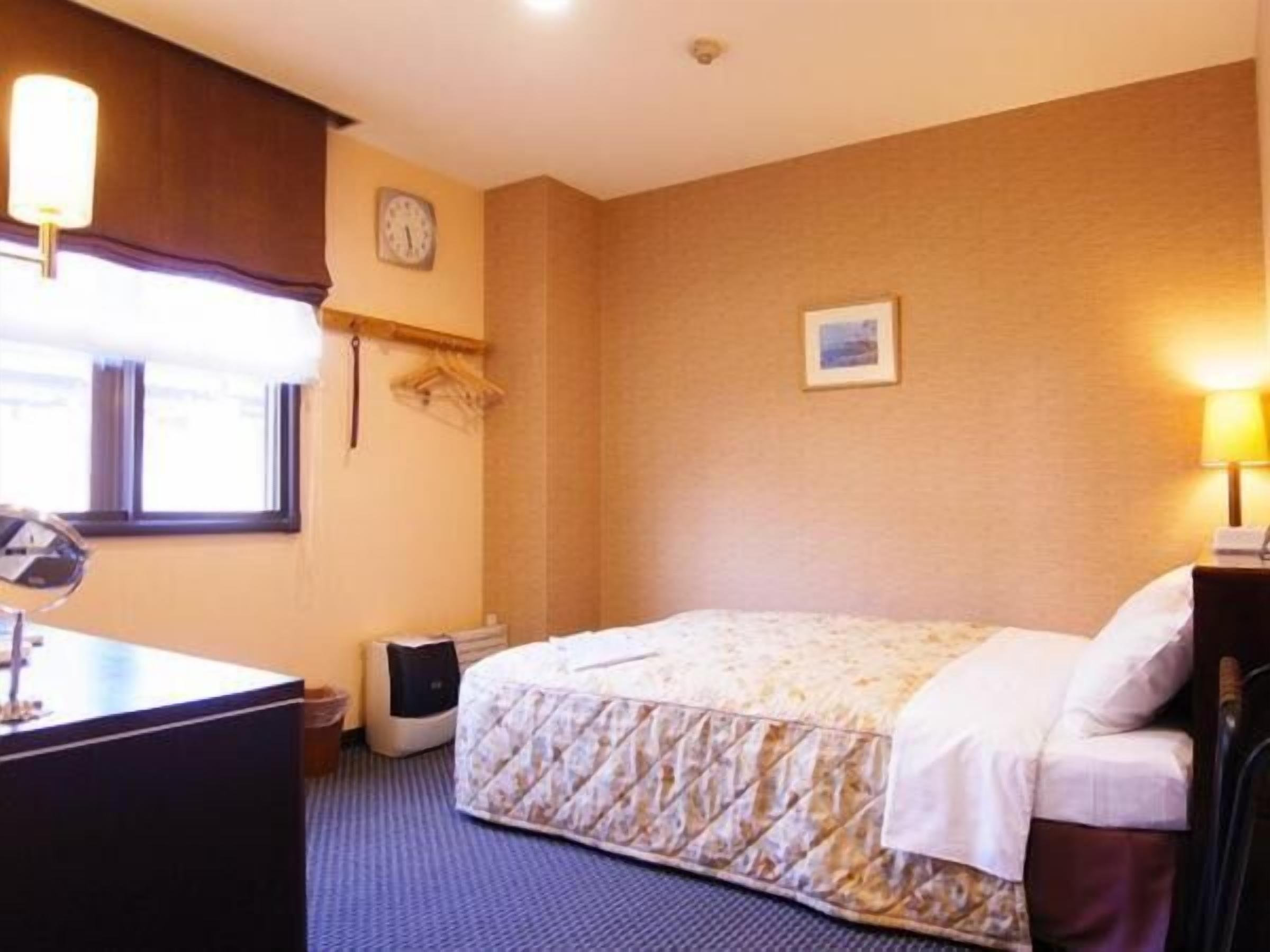 A hotel room with a bed, bedside table, lamp, clock, and a small picture on the wall.