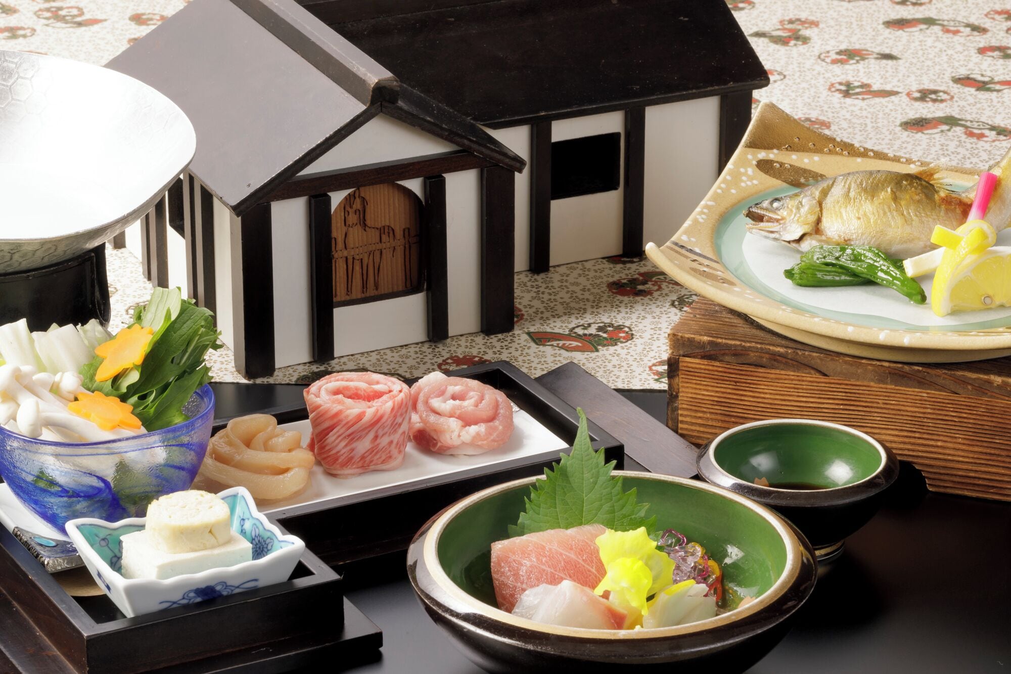 Una presentación tradicional de comida japonesa con diversos platos, incluyendo sashimi, verduras y un modelo decorativo de una casa.