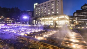 A hotel with multiple floors, a well-lit entrance, and a water feature in front. - Hotel Ichii (Kusatsu)