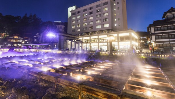 A hotel with multiple floors, a well-lit entrance, and a water feature in front. - Hotel Ichii (Kusatsu)