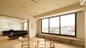 A spacious room with a large window offering a city view, a seating area with a sofa and chairs, and a flat-screen TV on a stand. - Hotel Ichii (Kusatsu)