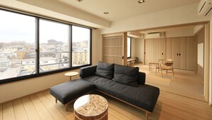 A modern living room with a large window, a sectional sofa, a small round table, and a view of the city. - Hotel Ichii (Kusatsu)