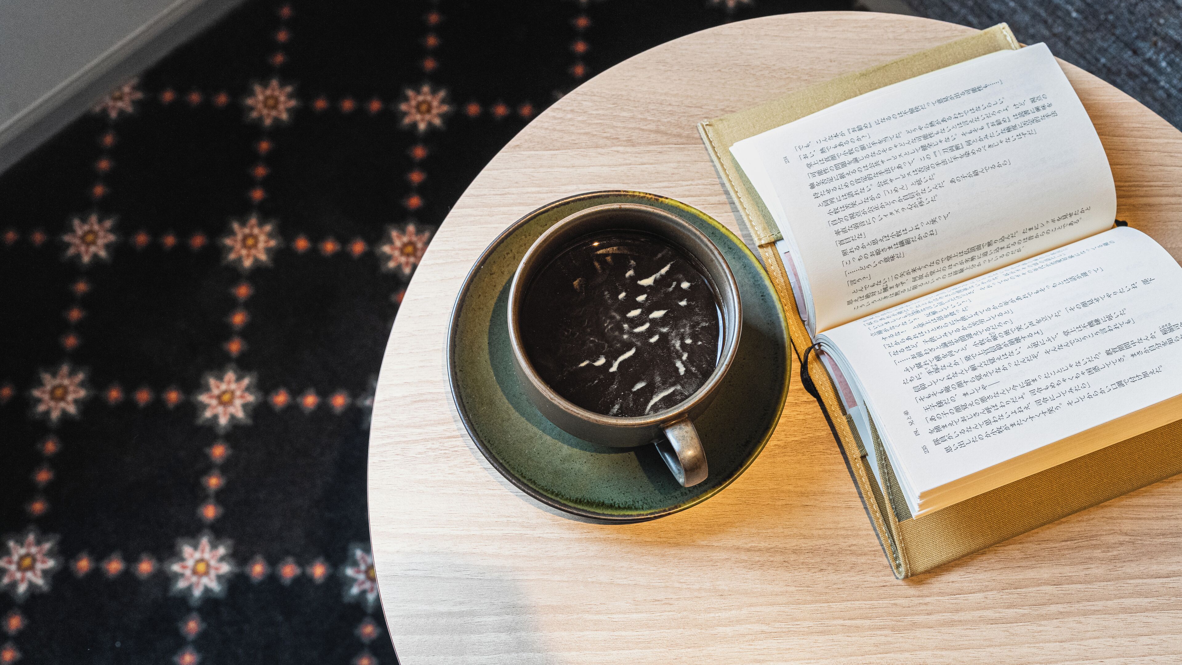 Eine Tasse schwarzer Kaffee auf einem runden Tisch, neben ihr ein offenes Buch.