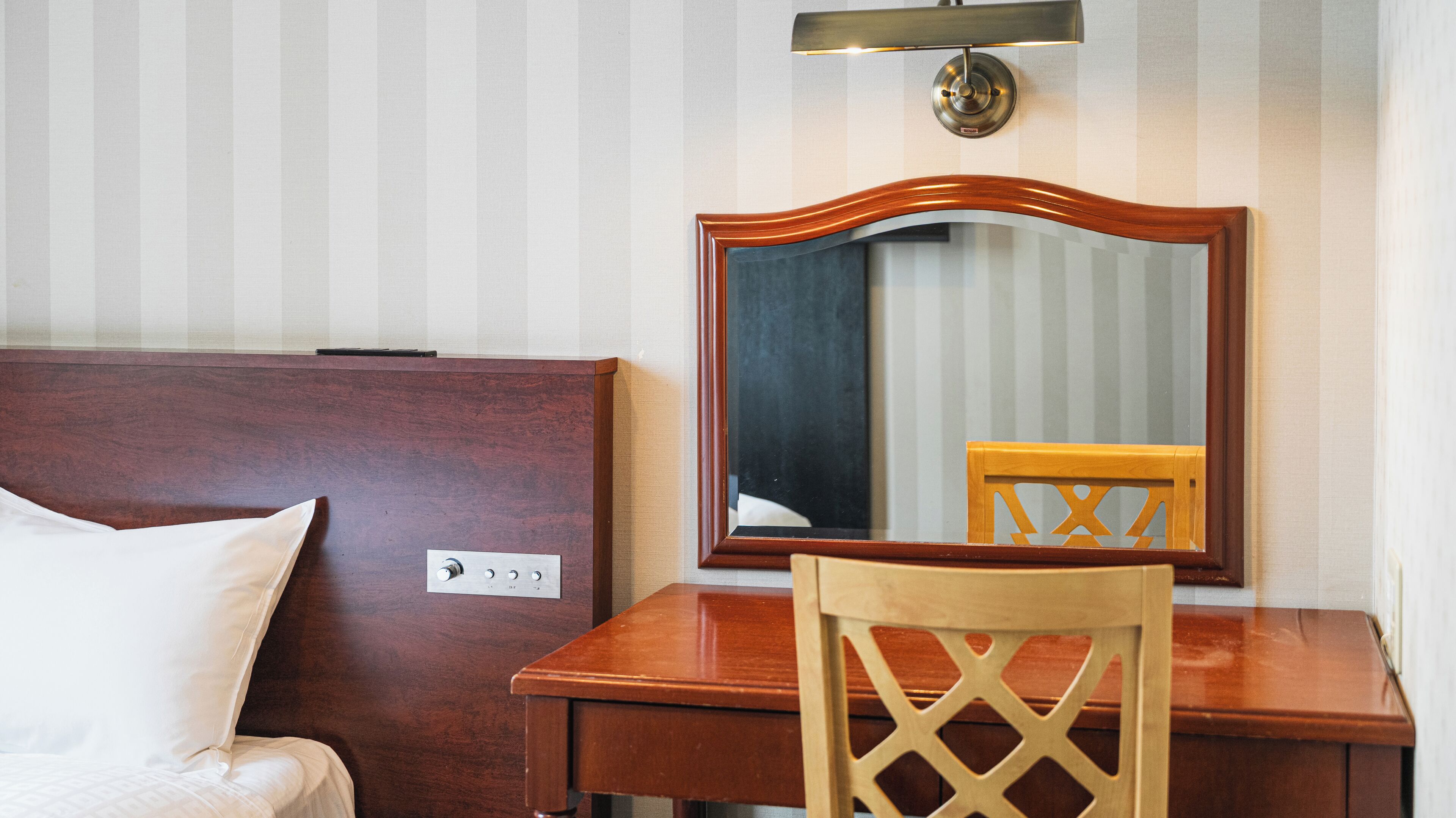 Habitación de hotel con cabecera de madera, mesita de noche, una silla, un espejo y una lámpara de pared.