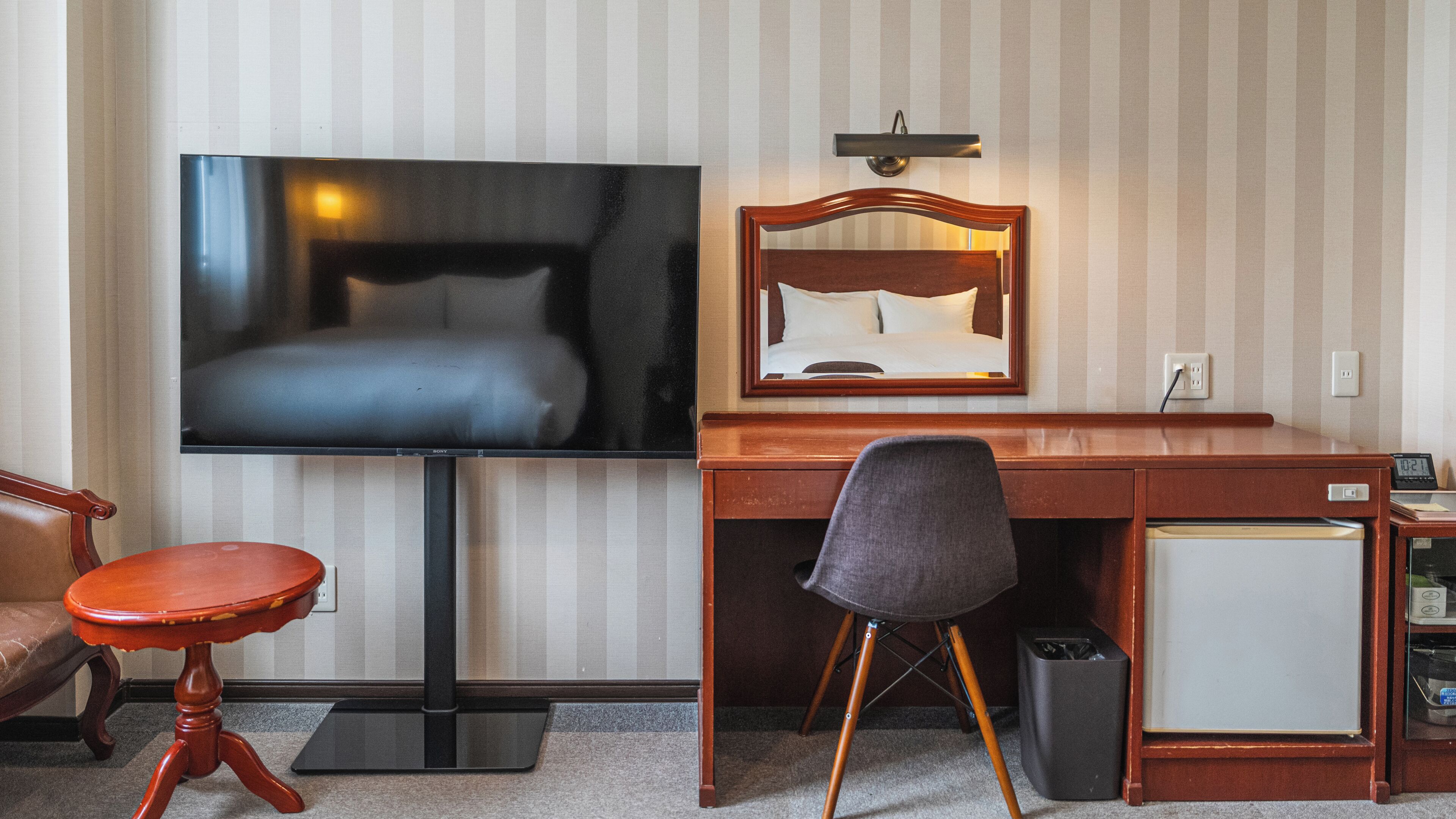 A hotel room with a bed, a TV, a desk, a chair, a small fridge, and a trash bin.