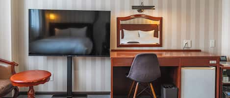 A hotel room with a bed, a TV, a desk, a chair, a small fridge, and a trash bin.