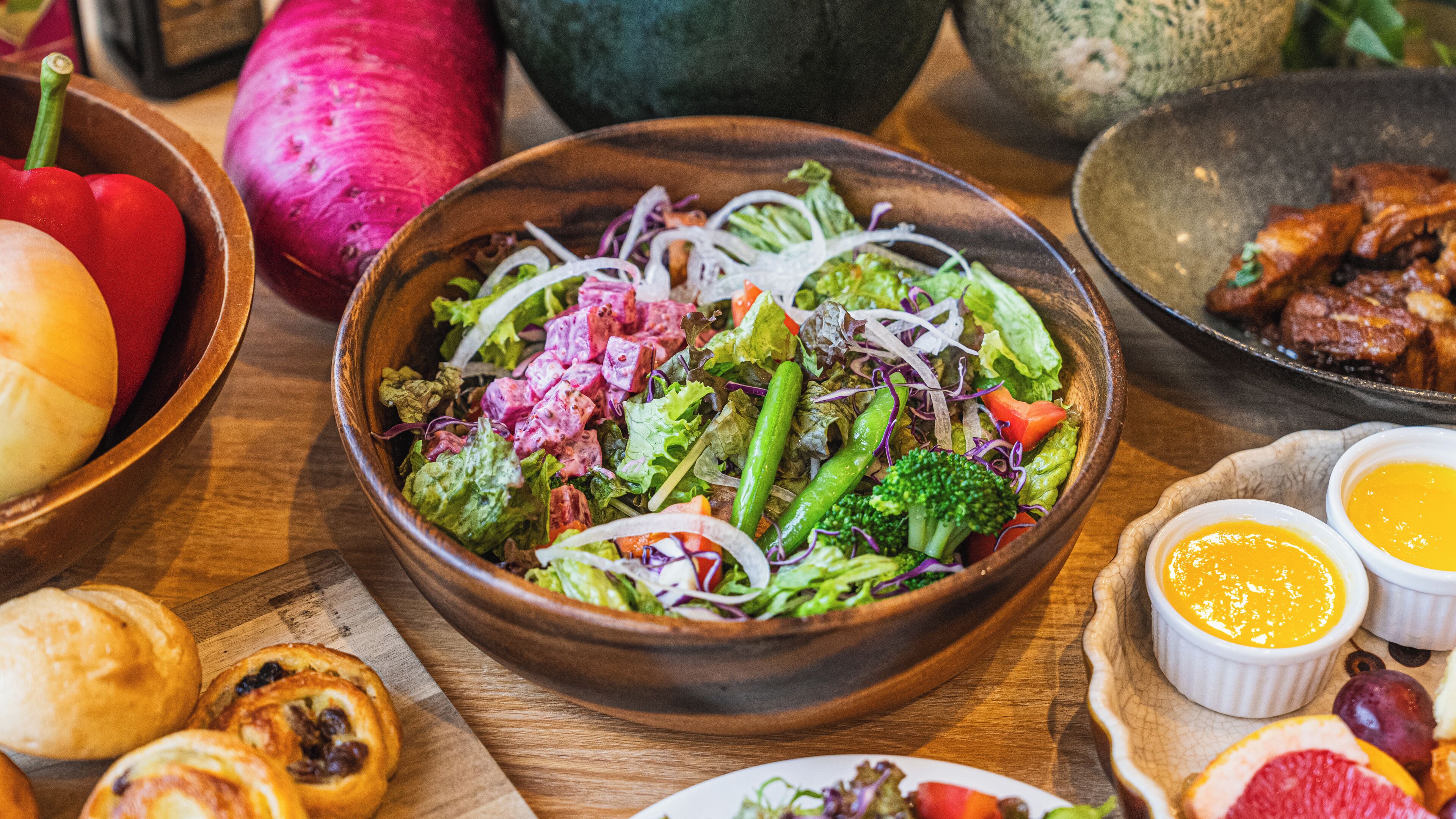 A wooden bowl filled with a salad, surrounded by various food items and bowls on a wooden table.