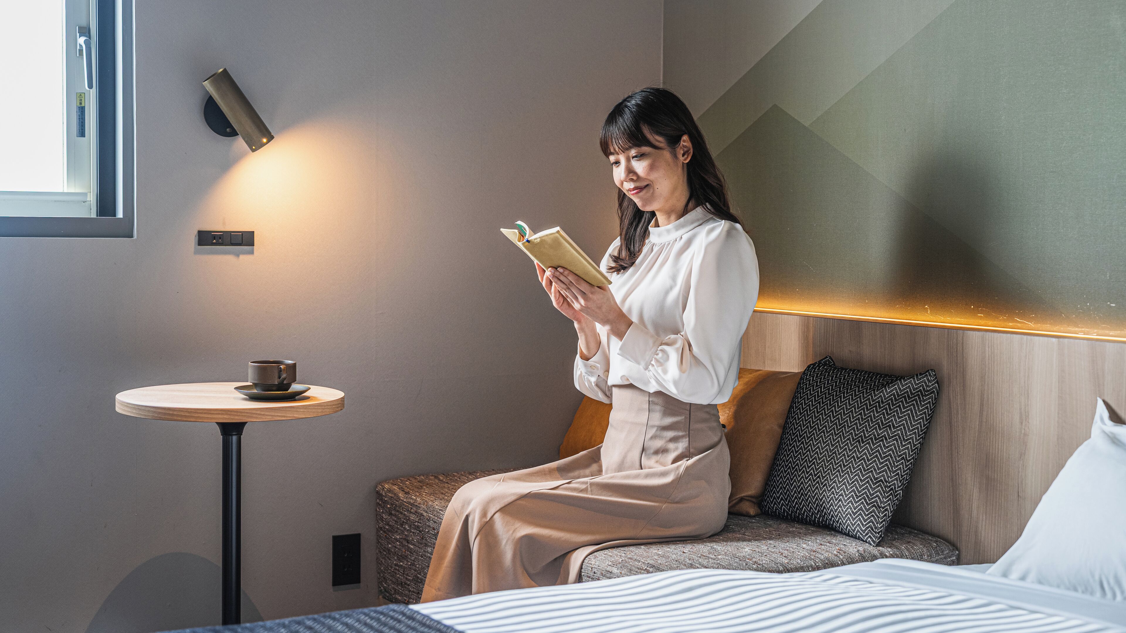 Een vrouw zit op een bed te lezen, met een klein tafeltje en kopje ernaast.