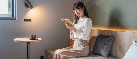 A woman sitting on a bed reading a book, with a small table and cup beside her.