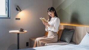 Una mujer sentada en una cama leyendo un libro, con una mesita y una taza cerca.