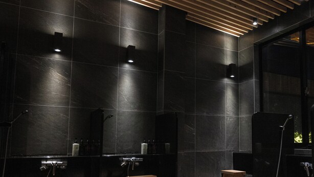 A modern bathroom with dark tiled walls, a wooden ceiling, and a large window. - Tsukuba No Yu Urban Hotel (Tsukuba)