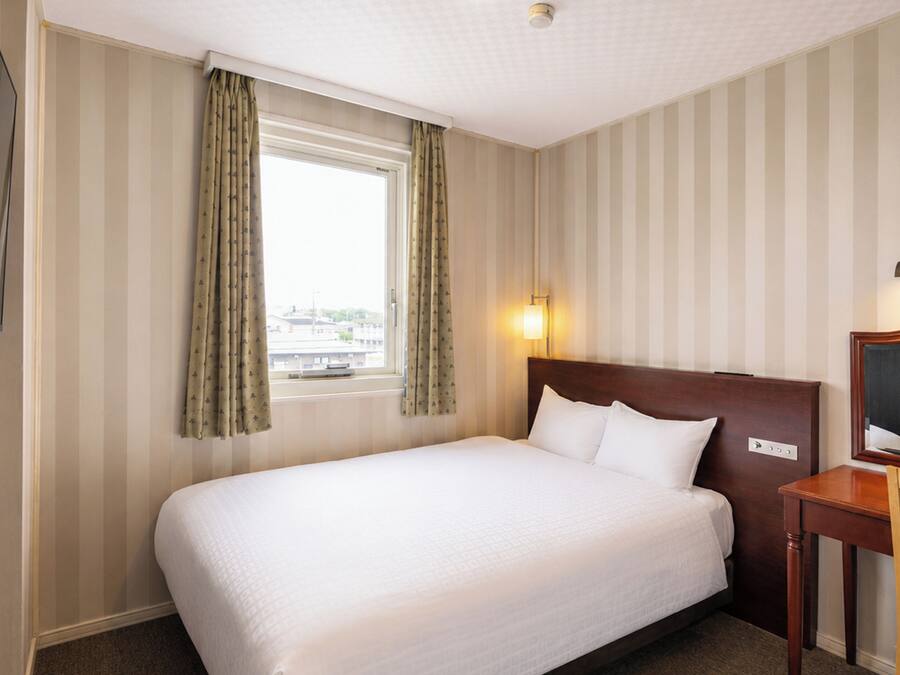 Una habitación de hotel con cama, escritorio, silla, televisor y ventana con cortinas.