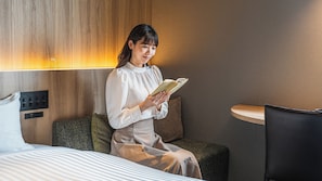 Una habitación de hotel con una cama, un sillón de lectura, una mesita de noche y un panel de control en la pared.