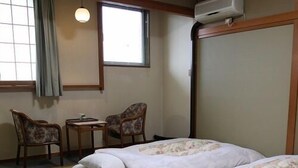 A hotel room with two beds, a small table, two chairs, a window with blinds, and an air conditioning unit. - Tsukuba Town Hotel (Joso)