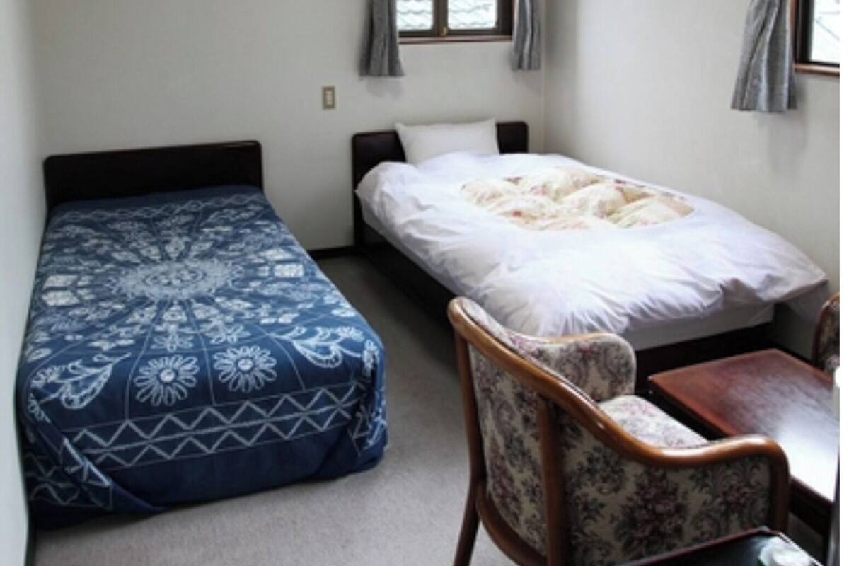Een kleine, eenvoudige kamer met twee bedden, een stoel en een tafel.