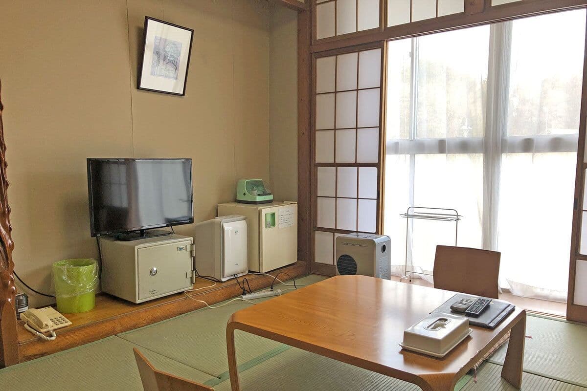 A traditional Japanese room with tatami flooring, a wooden table, a television, a small refrigerator, and a futon bed.