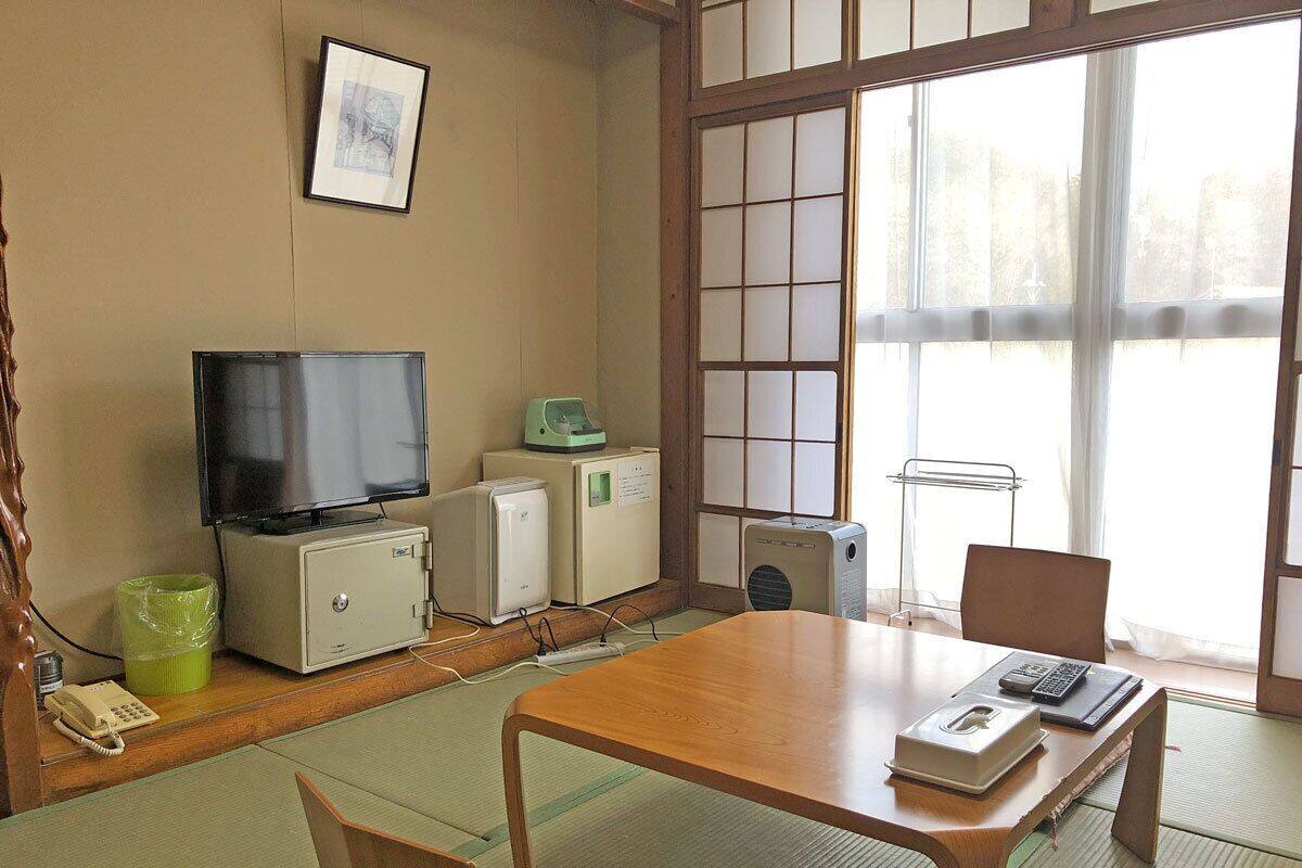 Una habitación japonesa tradicional con suelo de tatami, una mesa de madera, televisor, un refrigerador pequeño y una cama futón.