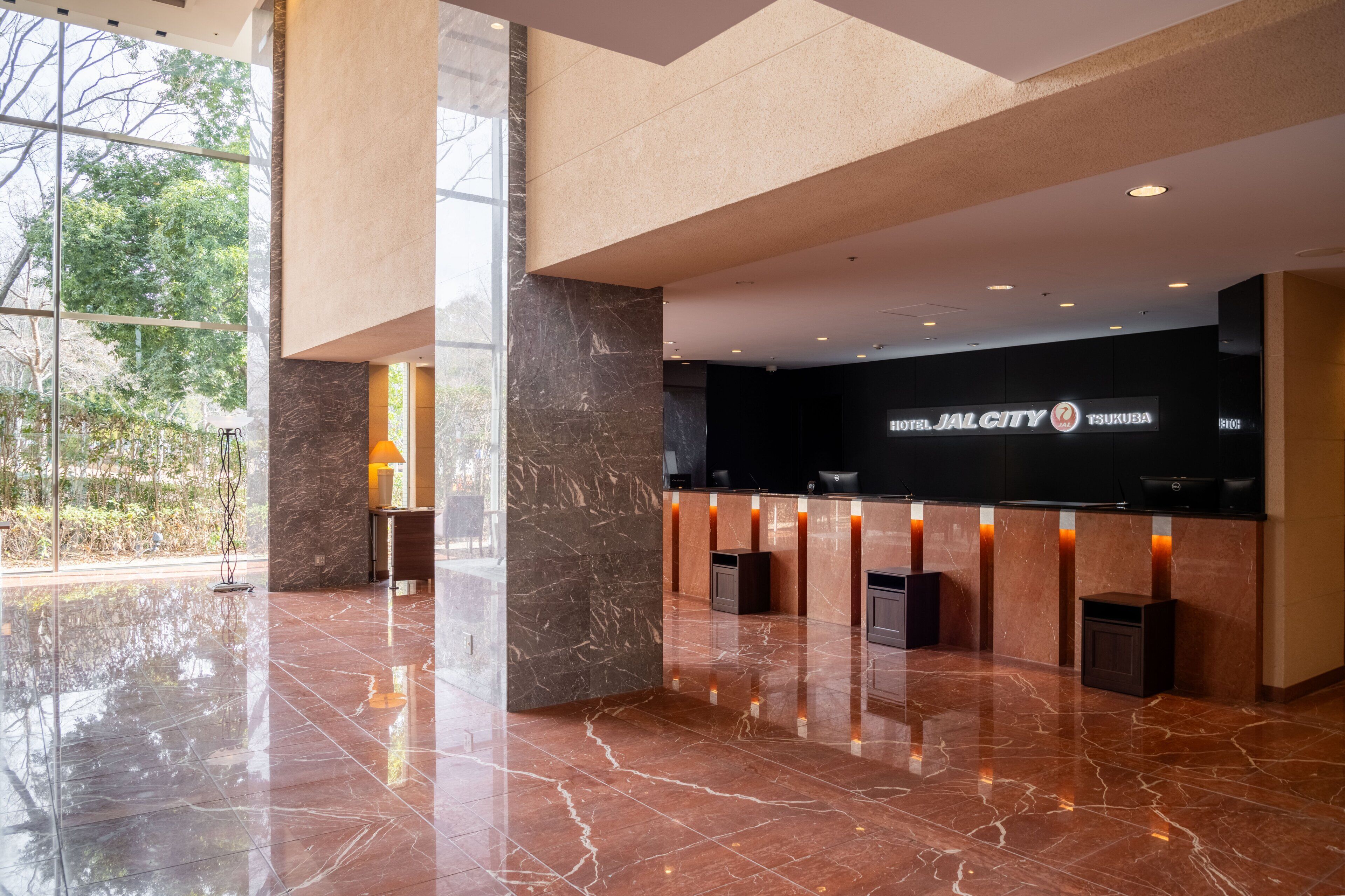 A modern hotel lobby with a reception desk, marble flooring, and large windows offering a view of the outdoors.