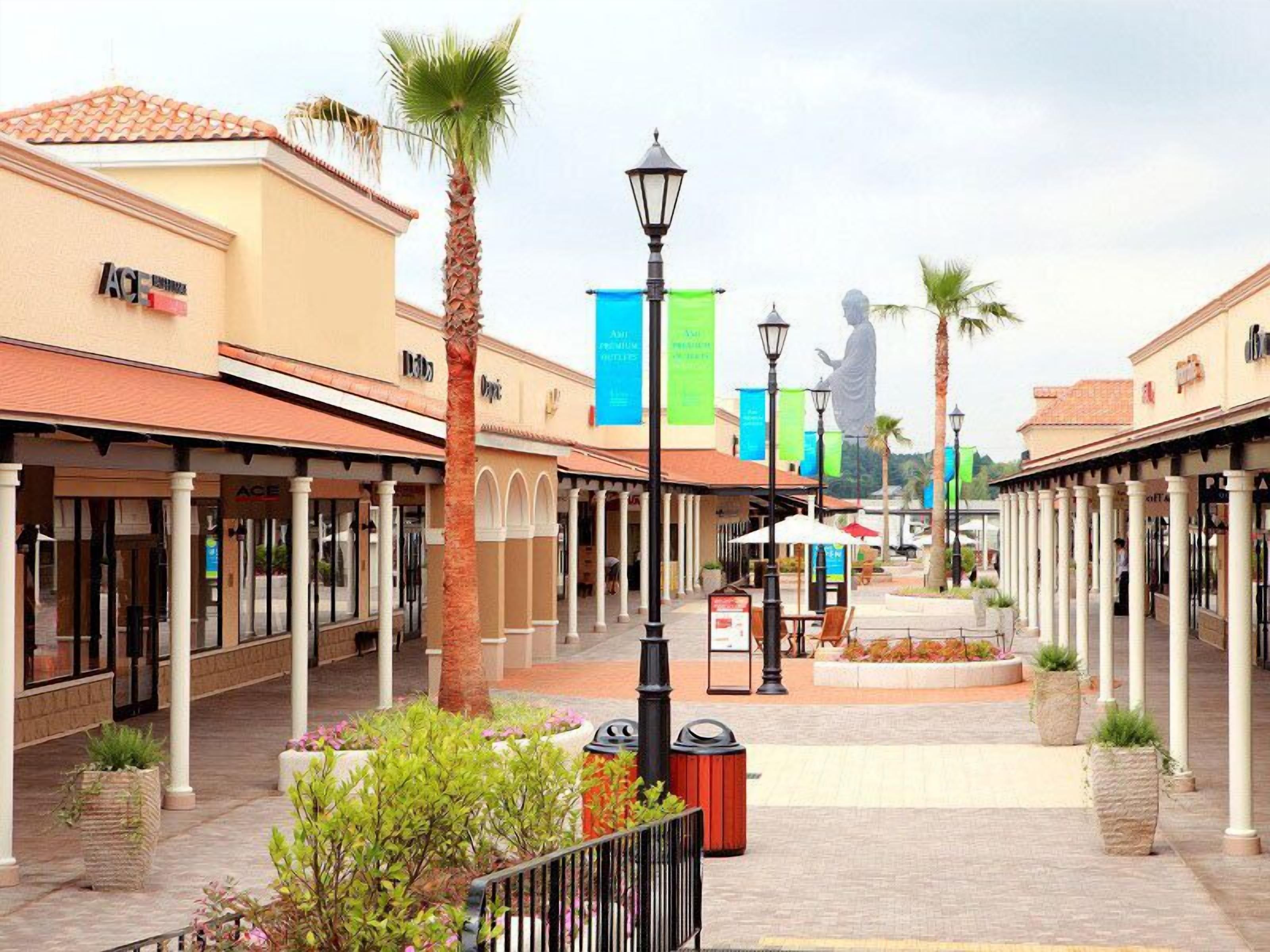 A shopping mall with covered walkways, palm trees, and a statue in the distance.