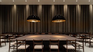 Un salón de conferencias con una gran mesa rectangular rodeada de sillas de madera con asientos acolchados, dos lámparas colgantes negras y un fondo de cortinas verticales.