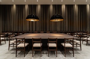 A conference room with a large rectangular table surrounded by wooden chairs with cushioned seats, two black pendant lights, and a backdrop of vertical drapery.