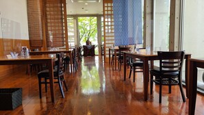 Un restaurante con mesas y sillas de madera, piso mojado y grandes ventanales con vistas a árboles.