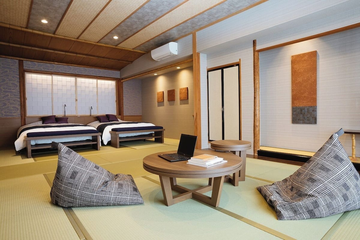 A traditional Japanese-style room with tatami flooring, two beds, a small round table with a laptop, and a futon.