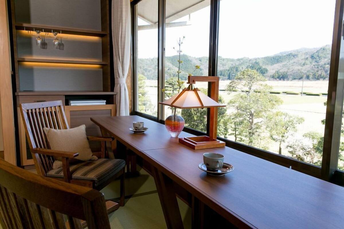 A room with a wooden table, chairs, a lamp, and a view of a landscape through large windows.
