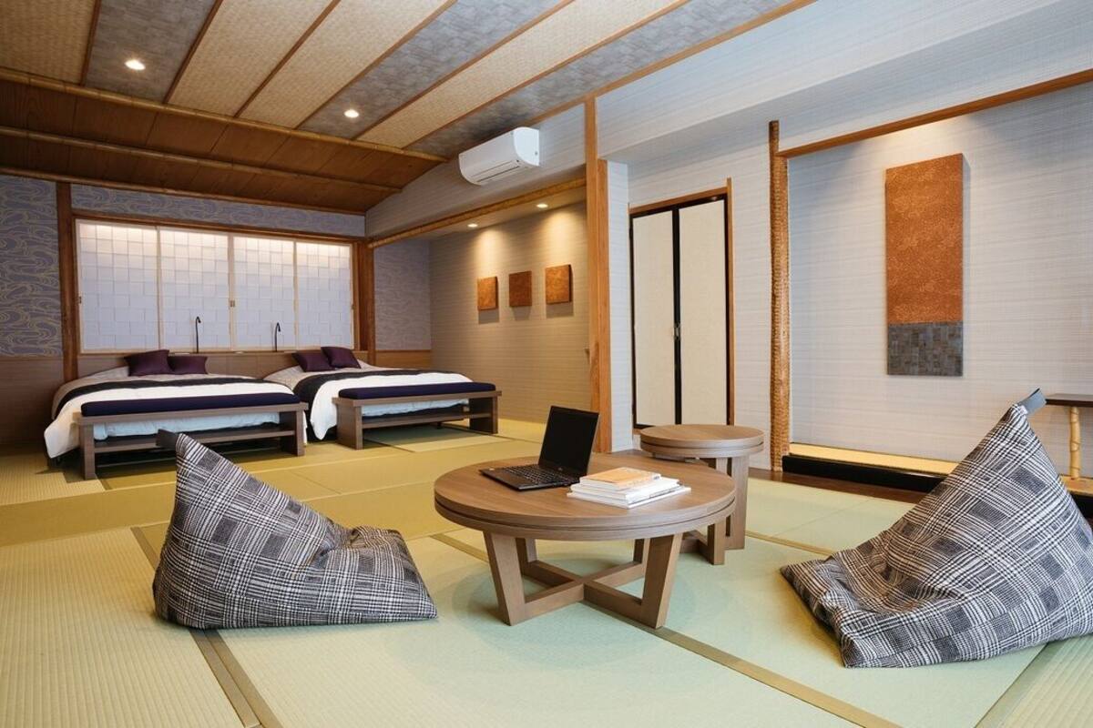 A traditional Japanese-style room with tatami flooring, two beds, a small round table with a laptop, and a wall with sliding doors.
