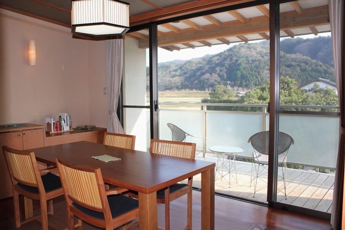 A dining area with a wooden table and chairs, a small kitchen area, and a balcony with a view of a lake and hills.
