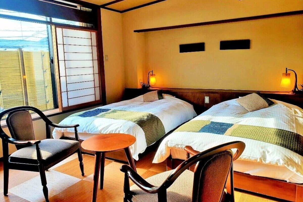 A hotel room with two beds, a small table, chairs, a window with blinds, and wall-mounted lamps.