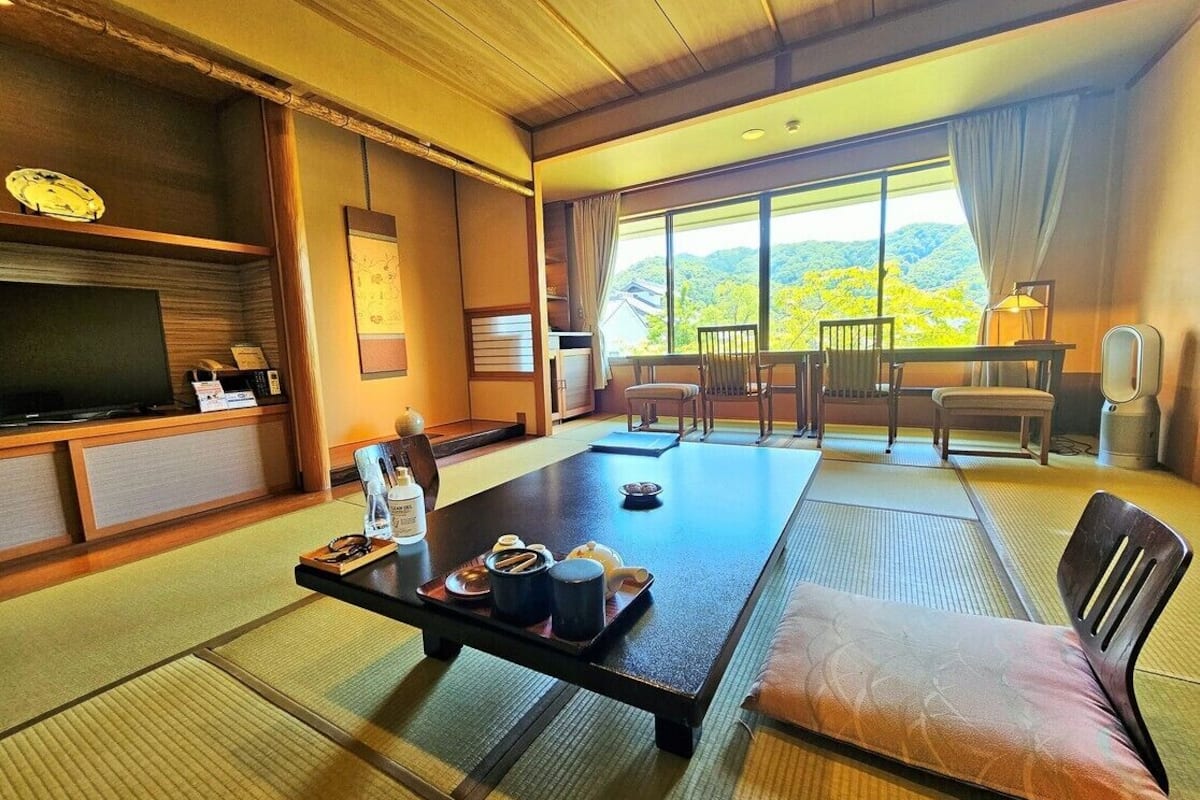 A traditional Japanese room with tatami flooring, a low table with a cushion, a television, and a view of the outdoors.