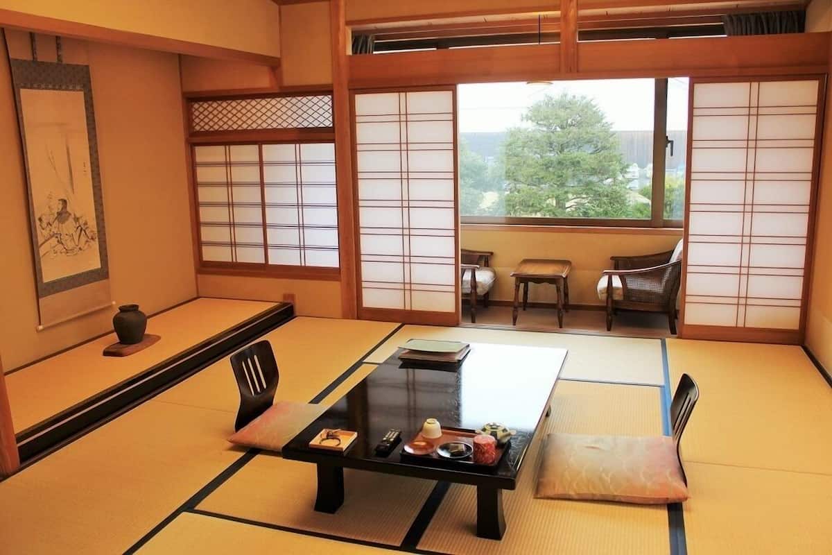 A traditional Japanese room with tatami flooring, a low table with a tray of items, sliding doors, and a view of greenery outside.
