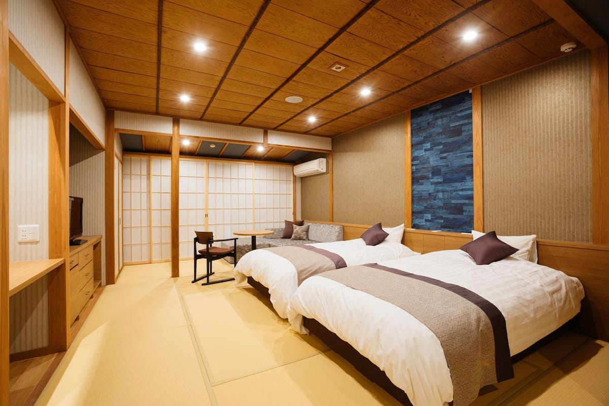 A traditional Japanese-style room with tatami flooring, two beds with pillows, a small table, and a TV.
