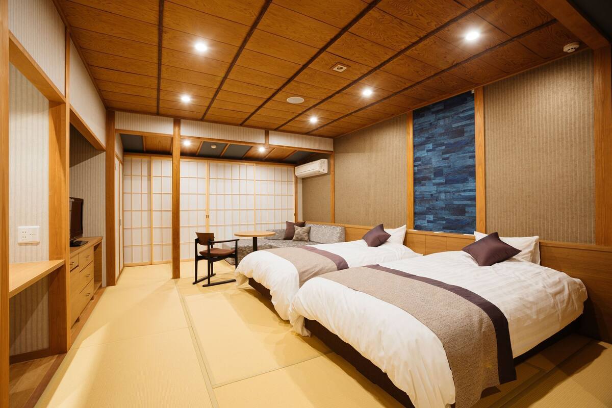 A traditional Japanese-style room with tatami flooring, two beds with pillows, a small table, and a TV.