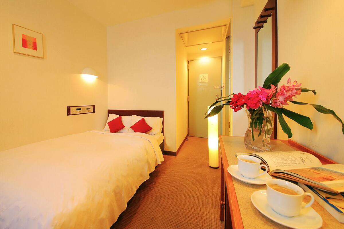 A hotel room with a single bed, a bedside table with a vase of flowers, a cup of coffee, and a book.